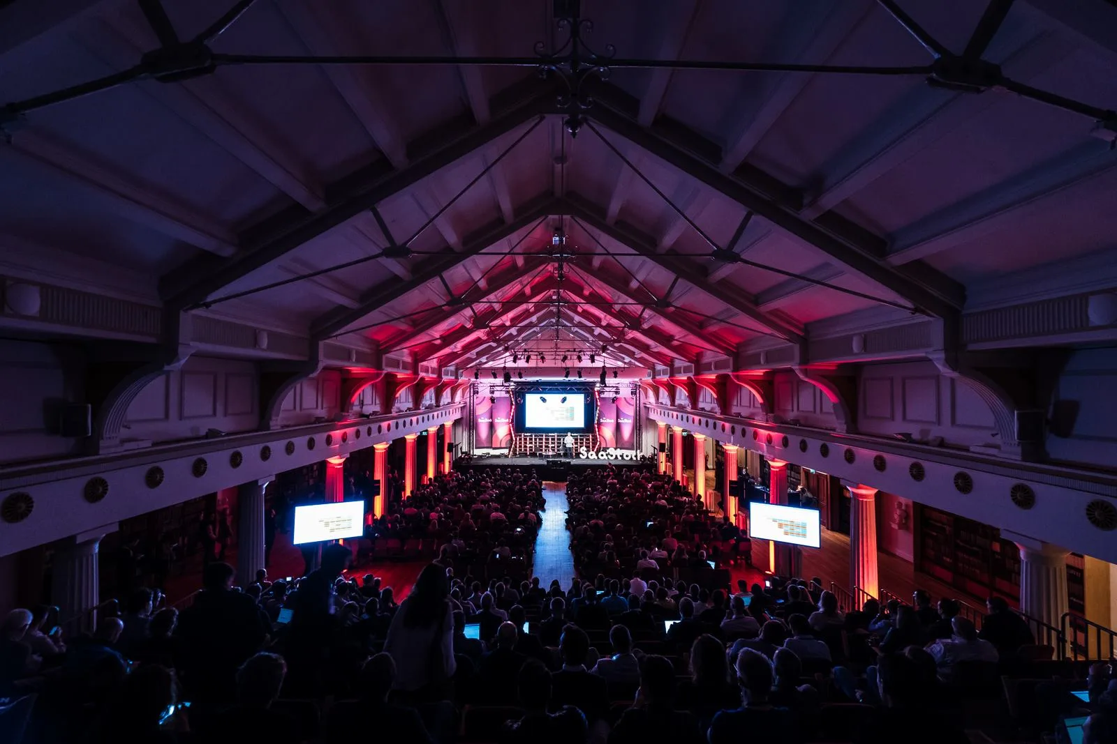 Live at SaaStock Europe 2017 — SaaStock mainstage opening from the balcony at RDS Industries Hall, Dublin, with red and pink uplighting along the original Victorian timber-vaulted roof and every row filled