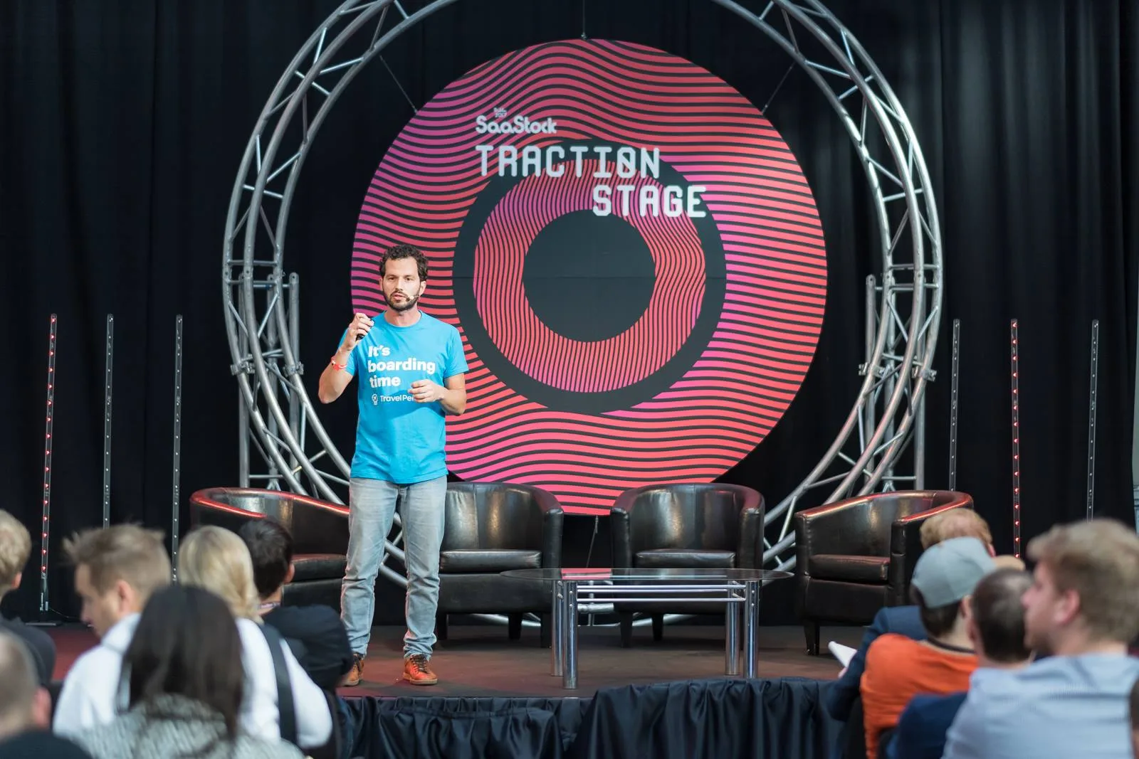 Live at SaaStock Europe 2017 — a TravelPerk speaker in the 'It's boarding time' tee mid-talk on the SaaStock Traction Stage in front of the red SaaStock circular backdrop