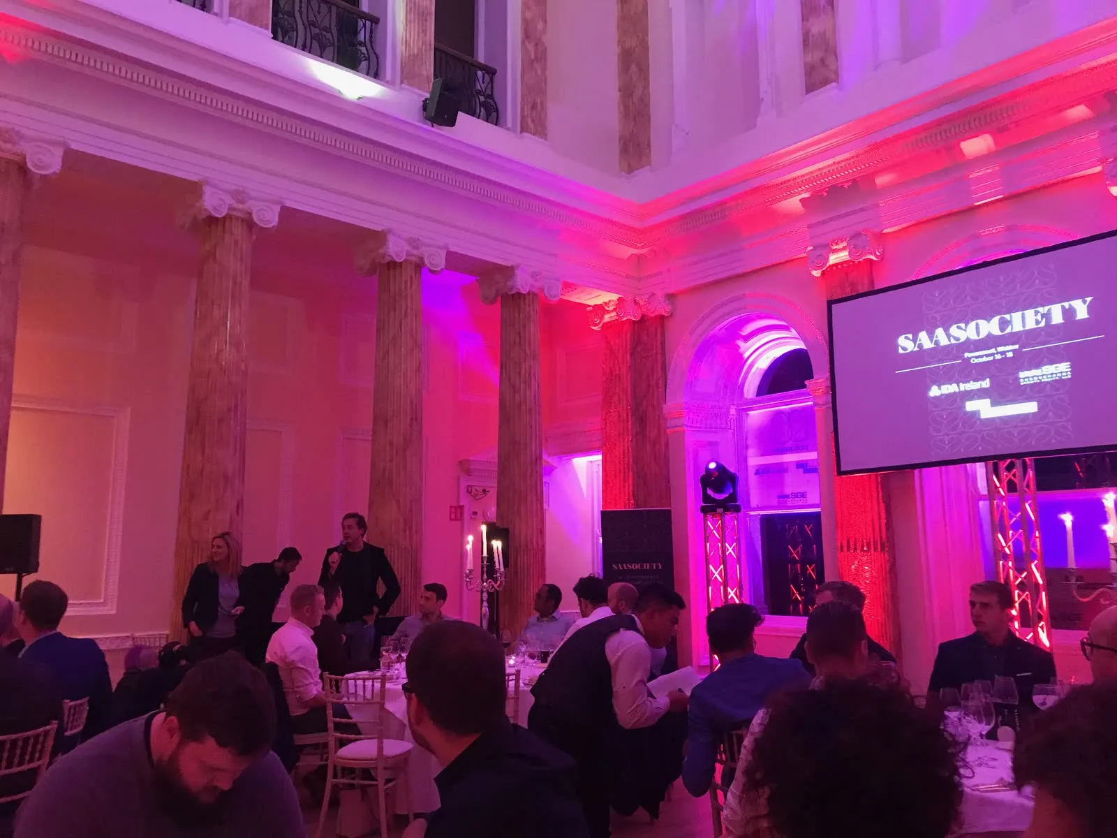 SaaSociety Founder Retreat 2019 — gala dinner in the columned, pink-uplit ballroom at Powerscourt Hotel Resort & Spa in Wicklow, founders seated at long candlelit tables under the SAASOCIETY title screen