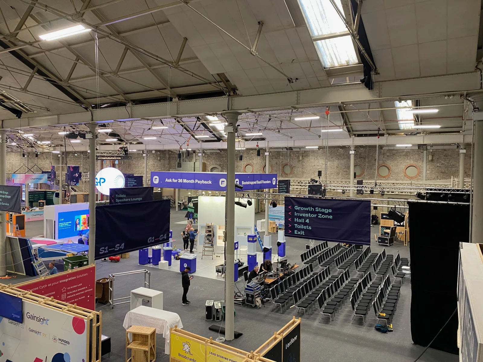 Pre-doors at SaaStock Europe 2022 — balcony overview of the expo hall mid-build with Founderpath '36 Month Payback' and 'Bootstrapped SaaS Founders' wayfinding banners hanging