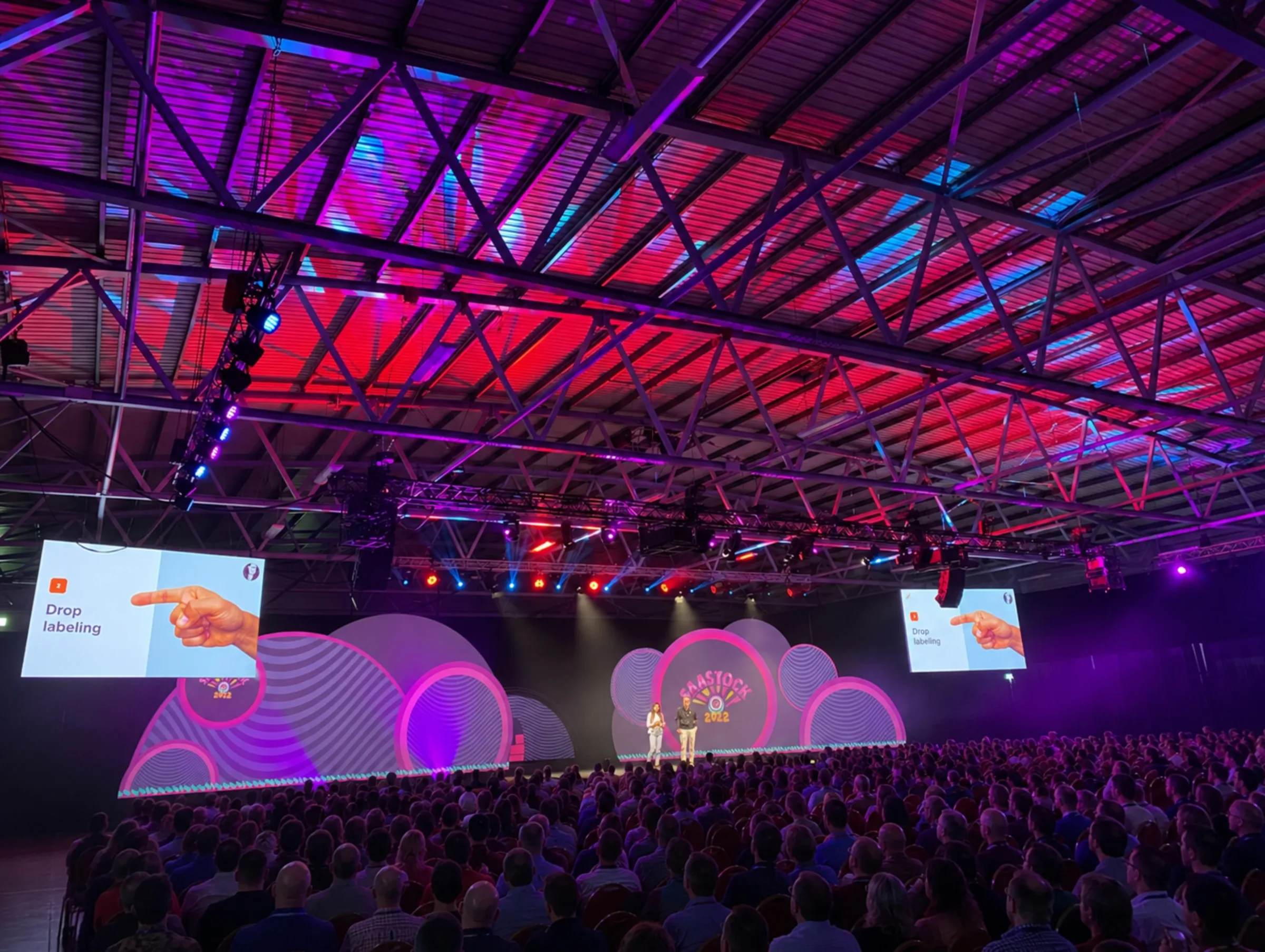 Live during SaaStock Europe 2022 — packed Mainstage under the red-and-pink ceiling wash, two presenters centre-stage in front of the cloud-shaped SaaStock 2022 LED set