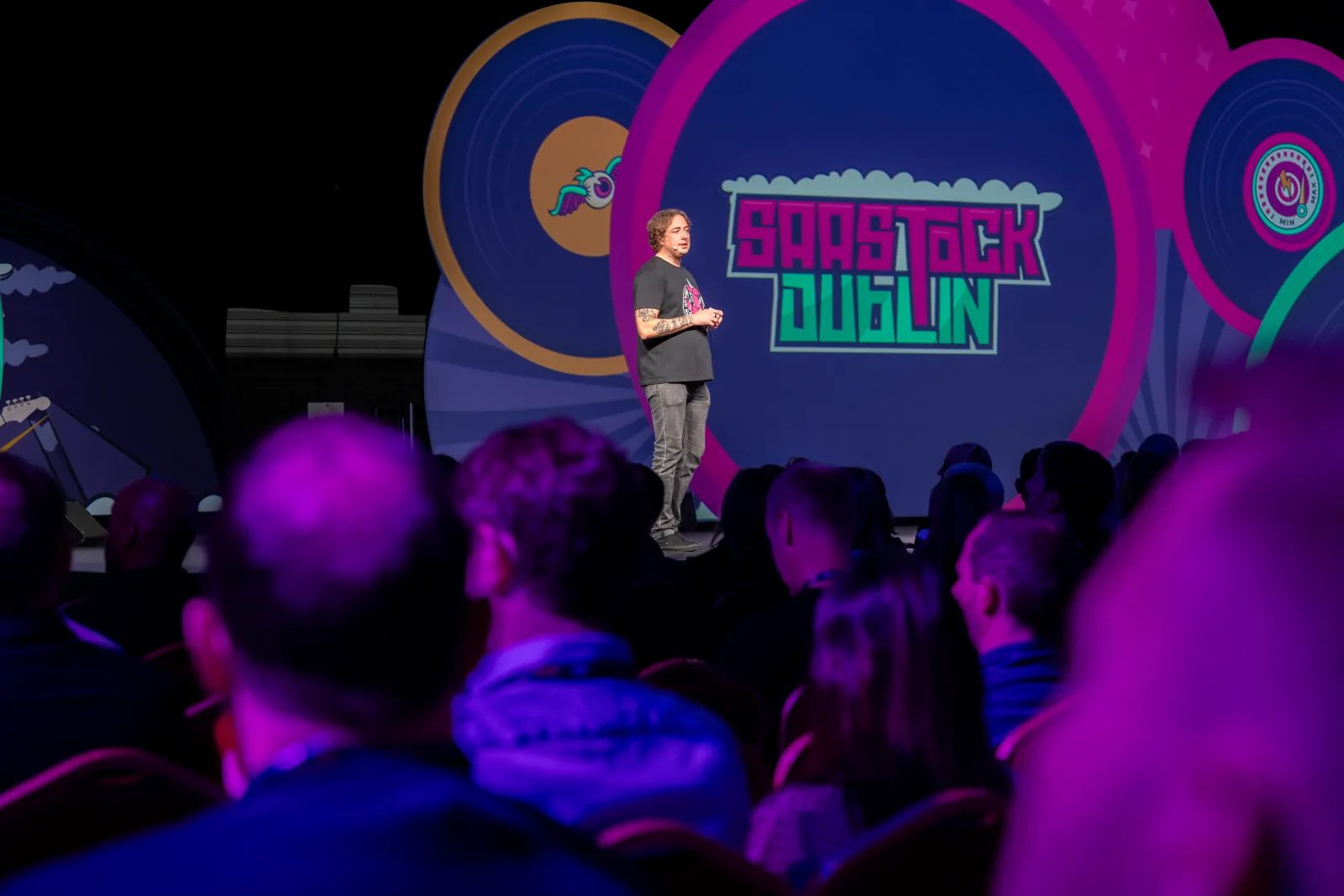 Mainstage keynote at SaaStock Europe 2023, RDS Simmonscourt, Dublin — speaker centered against the neon SaaStock Dublin LED backdrop with audience silhouettes in the foreground