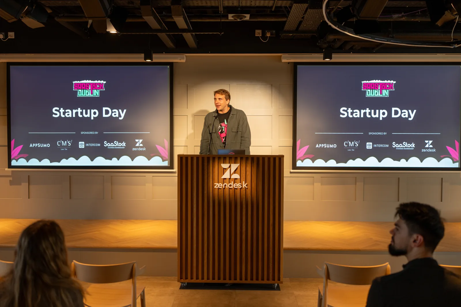Startup Day at the Zendesk Dublin office, Oct 15, 2023 — speaker behind the Zendesk podium with twin 'Startup Day' slides showing AppSumo, CMS Hub, Intercom and Zendesk sponsor logos