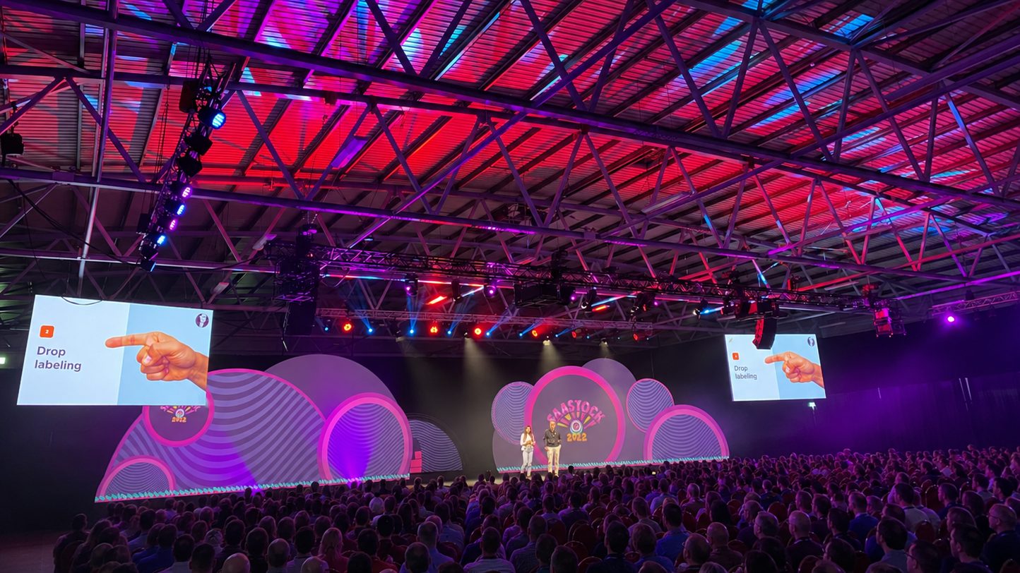 SaaStock Europe mainstage in Dublin — packed RDS arena with thousands of founders and operators in the audience, dual SaaStock keynote screens flanking the stage under magenta and purple lighting.