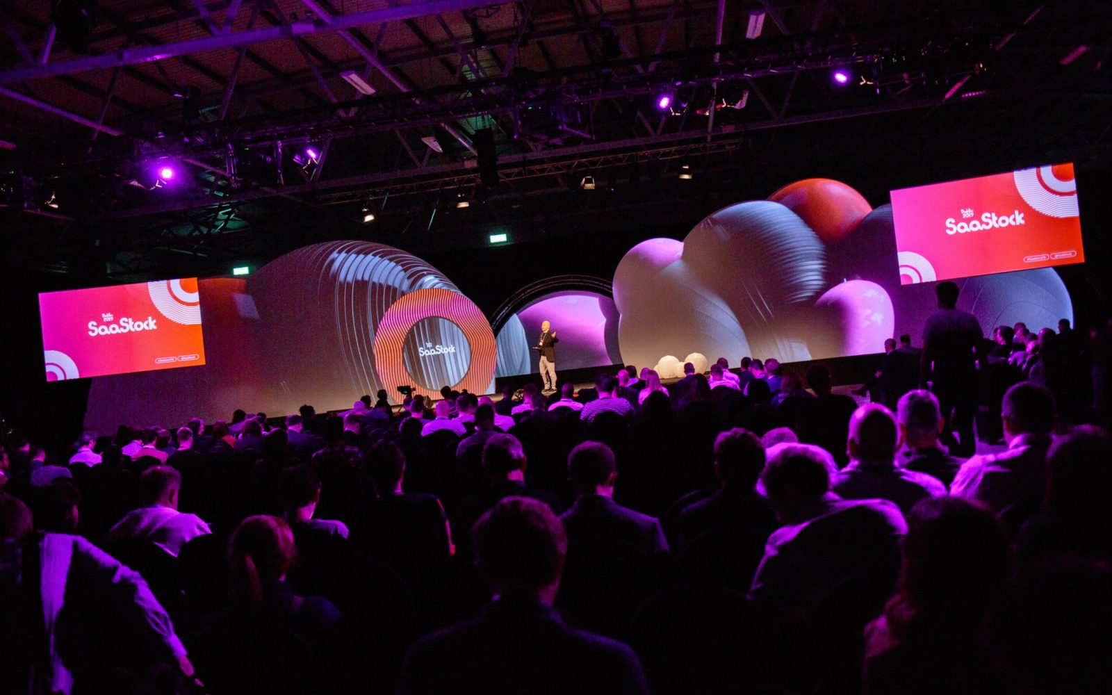 SaaStock mainstage in Dublin — a packed audience watching the SaaStock keynote with the brand-coloured sphere installations and dual SaaStock screens flanking the stage.