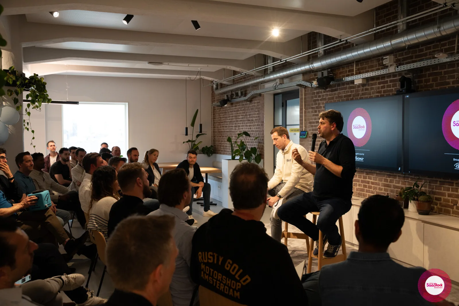 Live at SaaStock Local Amsterdam Q2 2024 — full room of ~35 Dutch SaaS founders seated in rows watching Ferdinand Goetzen and Joran Hofman on stage at Personio's Amsterdam office, with a 'Rusty Gold Motorshop Amsterdam' hoodie in the foreground and the SaaStock Local Amsterdam × Personio screens lit at the front