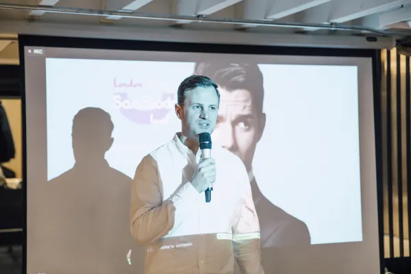 Live at SaaStock Local London — host at the microphone opening the evening in front of the projected SaaStock Local London title slide with a speaker headshot